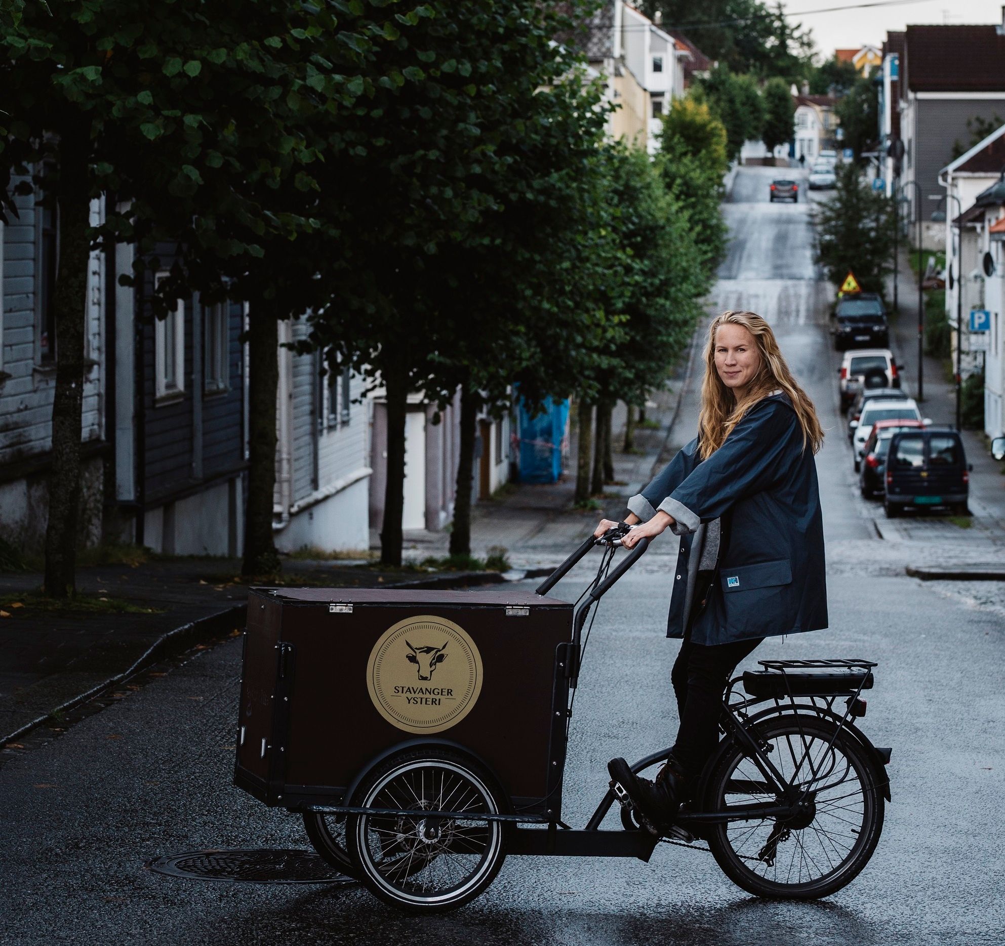 En person sykler på en gate med trær og hus på begge sider, og en lastebil med logo foran sykkelen.