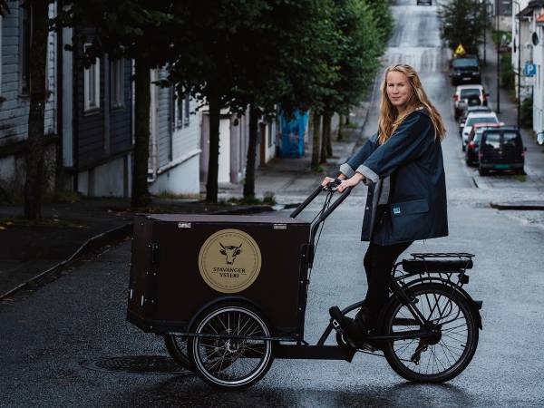 En person sykler på en gate med trær og hus på begge sider, og en lastebil med logo foran sykkelen.