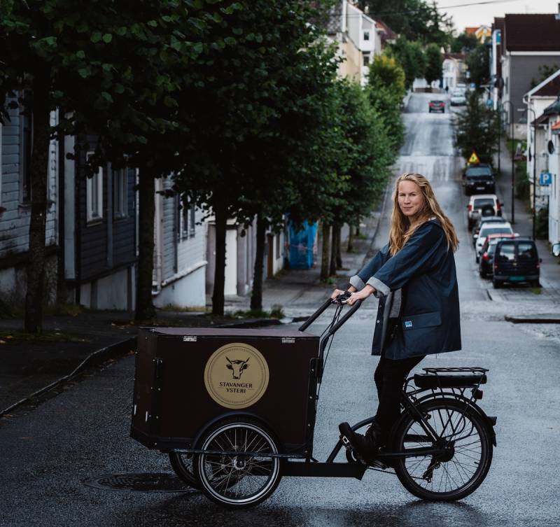 En person sykler på en gate med trær og hus på begge sider, og en lastebil med logo foran sykkelen.