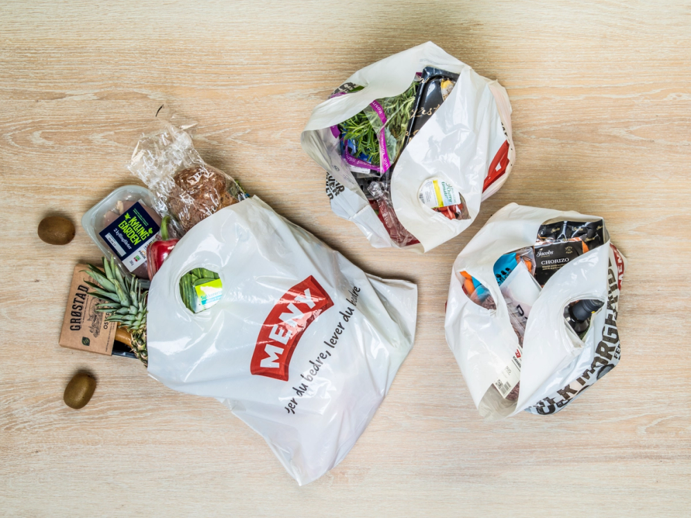 Poser med ulike aviser og reklamebrosjyrer på et trebord, med en plastpose fra Maxbo og noen nøtter ved siden av.