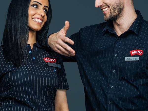 To ansatte i uniform med navnemerker står sammen og smiler, noe som kan symbolisere inkludering og mangfold på arbeidsplassen.