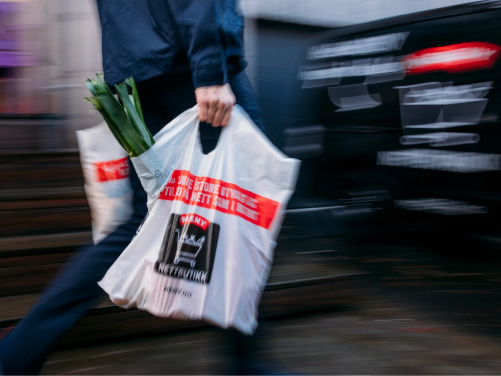 En person går med en stor hvit plastpose med tekst og logo, og en bil kjører forbi i bakgrunnen.