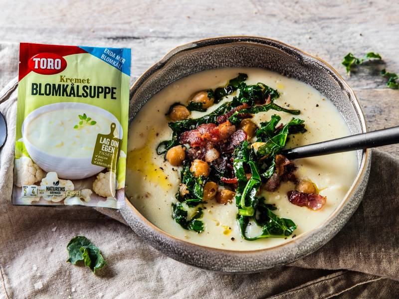 En skål med kremet blomkålsuppe med grønne grønnsaker og baconbiter, plassert på et treverkbord med en skje i suppen, og en pose med Toro blomkålsuppe ved siden av.
