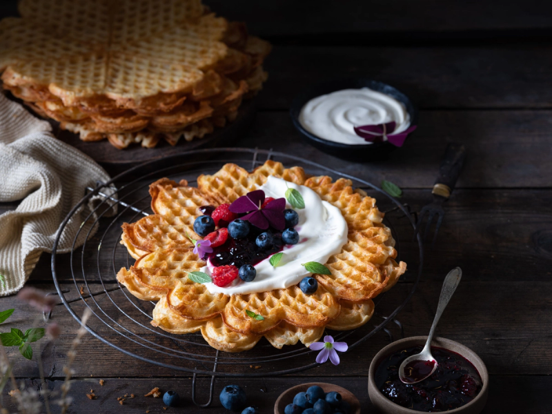 Vafler med rømme, bær og syltetøy på et svart bord med en skje og en liten skål med syltetøy ved siden av.