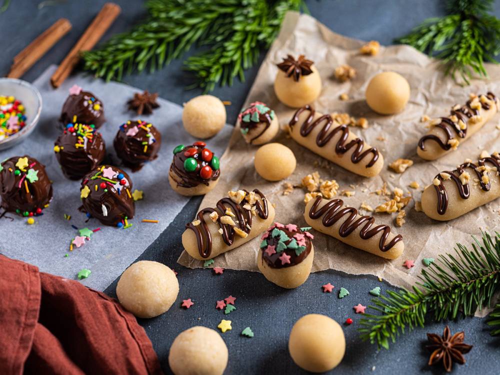 Hjemmelaget marsipan og sjokoladekonfekt pyntet med stjerner og småkaker på et bord med grønne grener rundt.