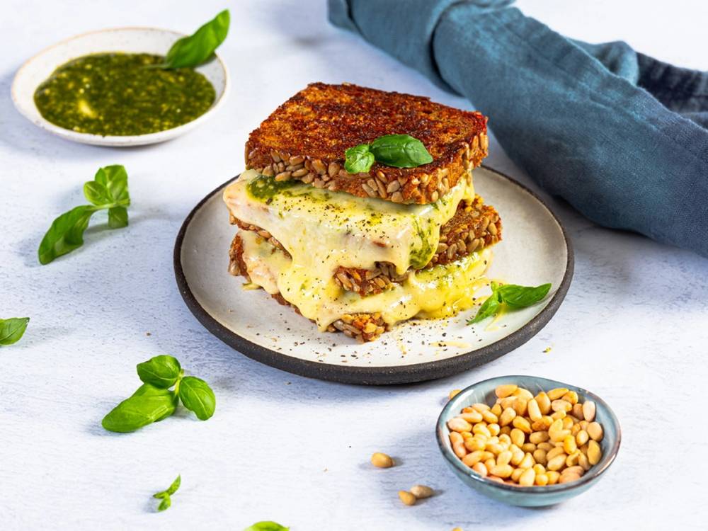 Et stykke toast med ost og grønnsaker på en tallerken, omgitt av basilikumblader og en liten skål med pinjekjerner, med en person i bakgrunnen som holder toasten.