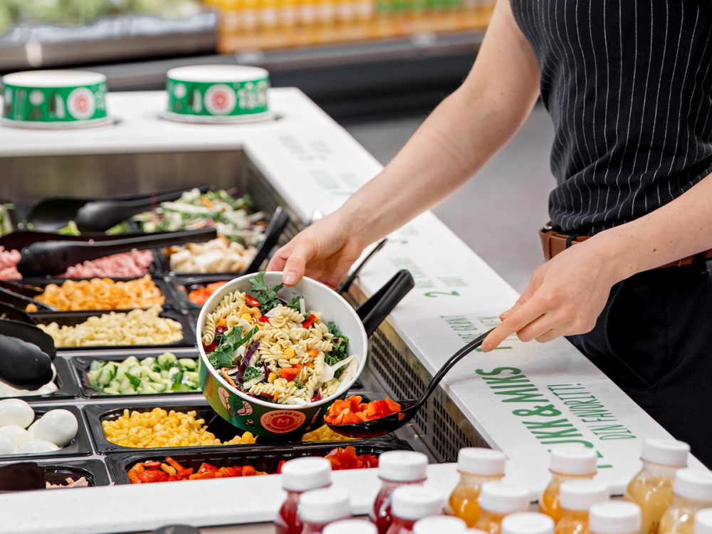 En person fyller en skål med salat fra en salatbar med ulike grønnsaker og tilbehør.