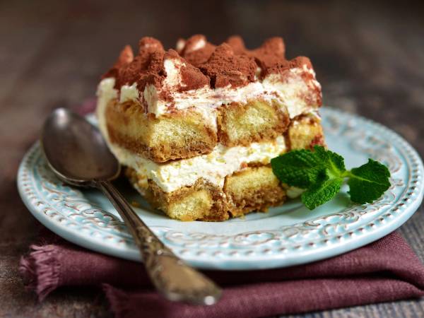 Et stykke tiramisu med lag av krem og kakaopulver på toppen, servert på en blå tallerken med en skje ved siden av.