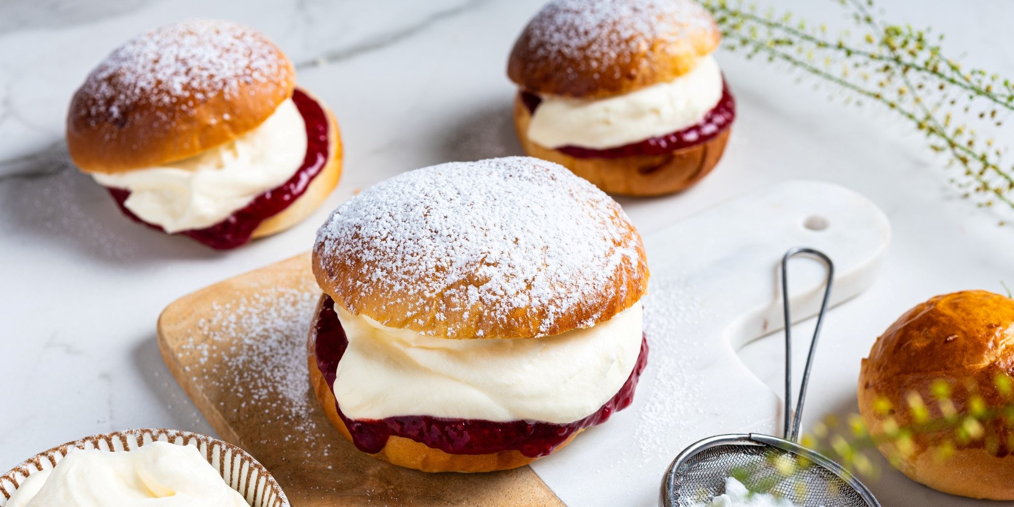 Tre fastelavnsboller med krem og syltetøy på et fat, pyntet med melis og omgitt av andre bakverk.