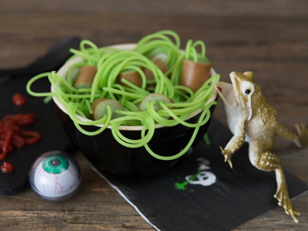 En skål med grønn spagetti-lignende pasta og en liten dinosaurfigur ved siden av på et svart underlag.