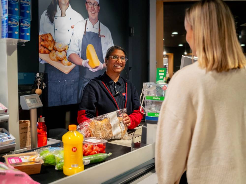 Kunde i kasseområdet på MENY Løren handler med en ansatt bak disken som har en plakat med bilder av matvarer i bakgrunnen.