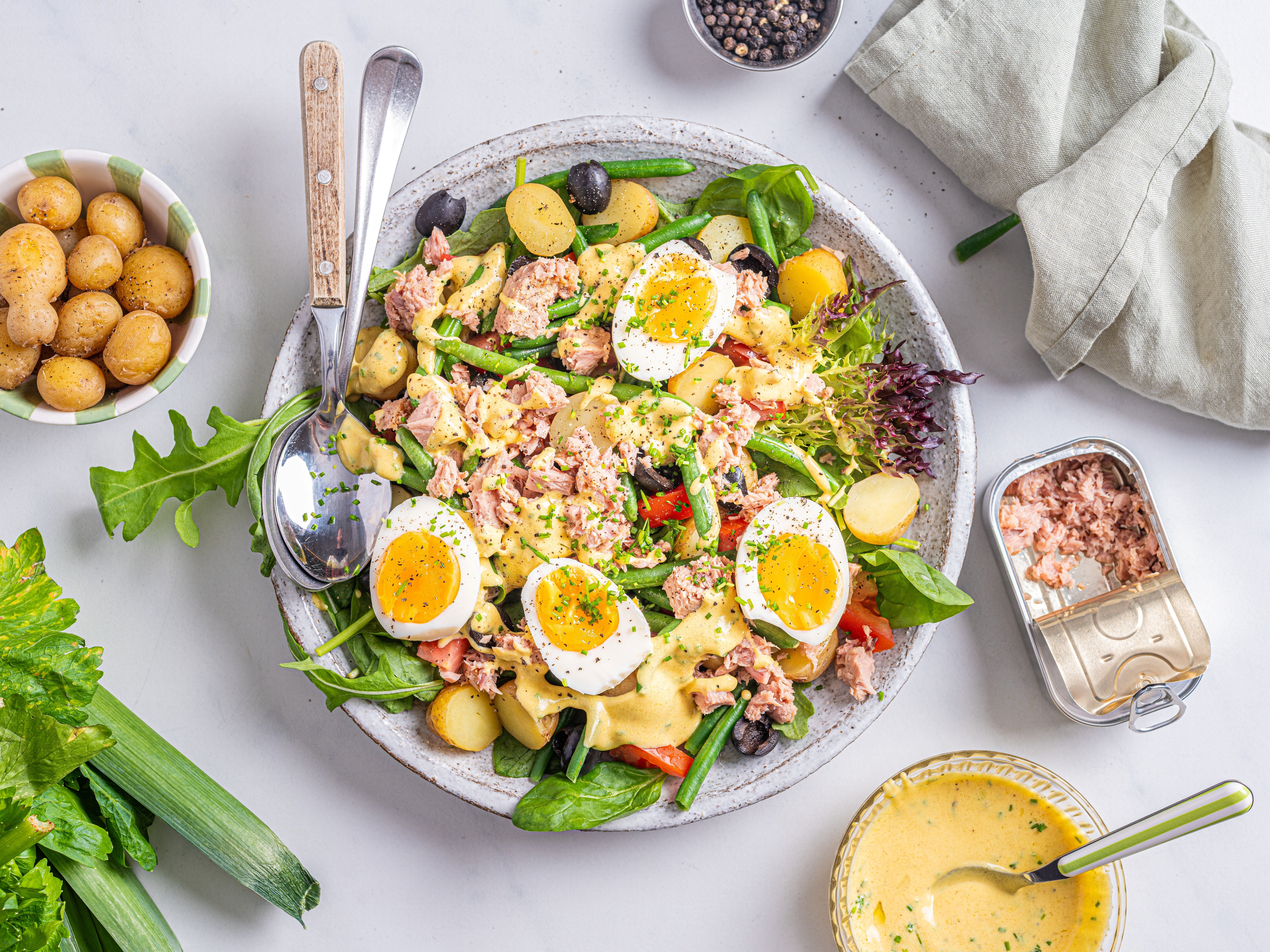 En skål med Nicoise-salat som inneholder egg, poteter, grønne bønner, tomater og tunfisk, omgitt av tilbehør som en liten skål med poteter og en bolle med dressing.