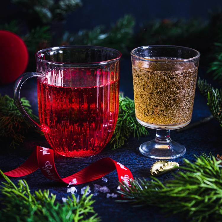 To glass med julebrus, ett i rød og ett i gullfarge, omgitt av julegrener og en rød julekule, på en mørk bakgrunn.