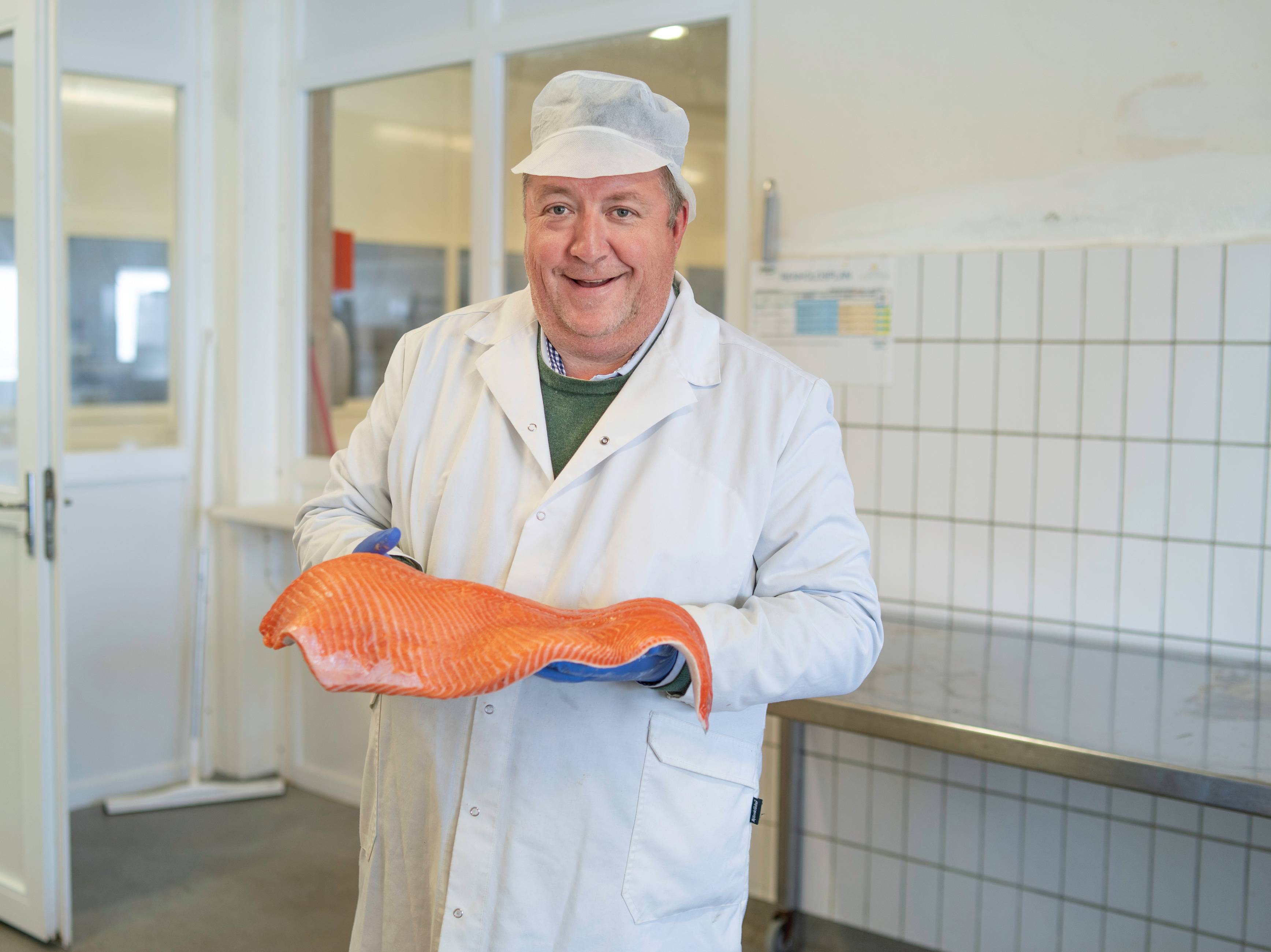 Person i hvit labfrakk holder en stor, oransje laksefilet i hendene på et fiskemottak eller kjøkken.