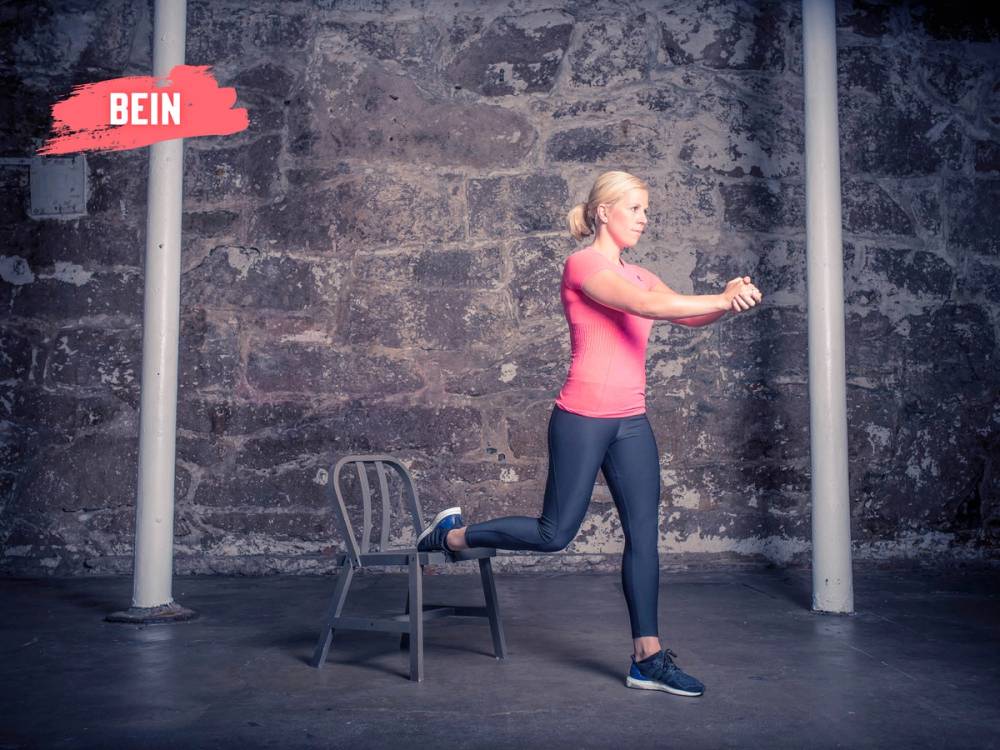 En person utfører en splitknebøy med en fot på en stol og den andre på gulvet foran en murvegg, med fokus på treningsøvelsen.