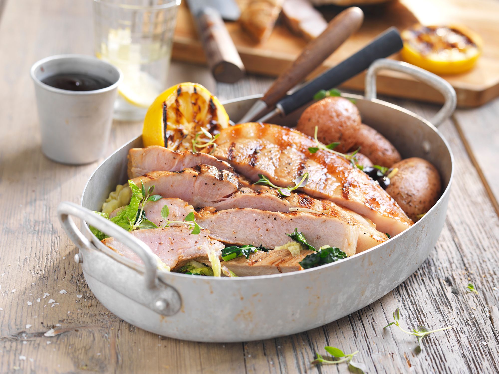 Flintstek med poteter og grønnsaker på et fat, omgitt av kjøkkenutstyr og en kopp med drikke.