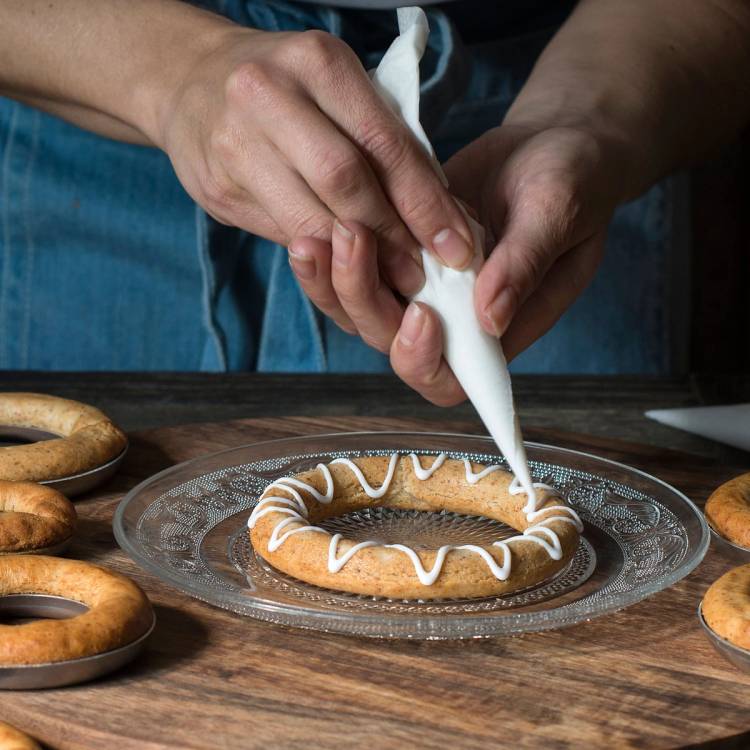 En person sprøyter hvit glasur på en kvadratisk kransekake med flere små kransekaker rundt på et bord.