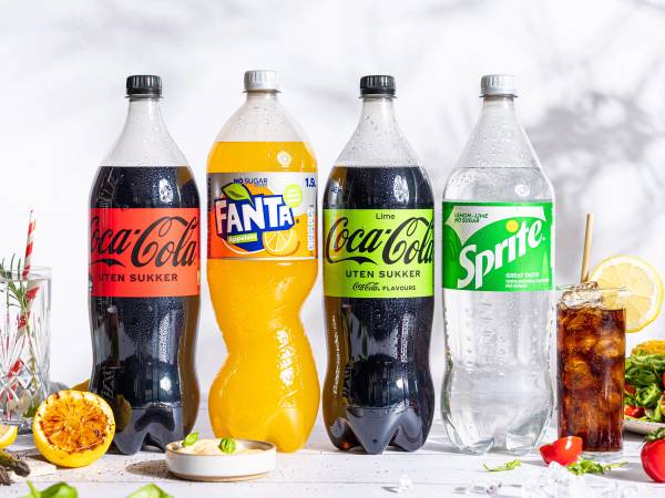 Fire brusflasker av Coca-Cola, Fanta og Sprite på et bord med noen frukter og isbiter, mot en lys bakgrunn.