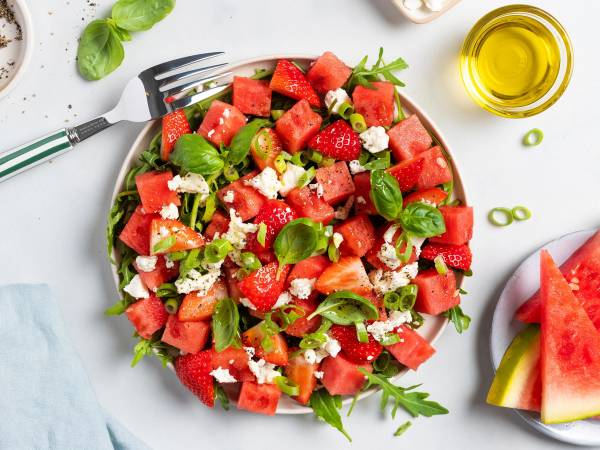 En frisk vannmelonsalat med jordbær, mynte og fetaost på en tallerken, omgitt av en skive vannmelon og en liten bolle med olje.