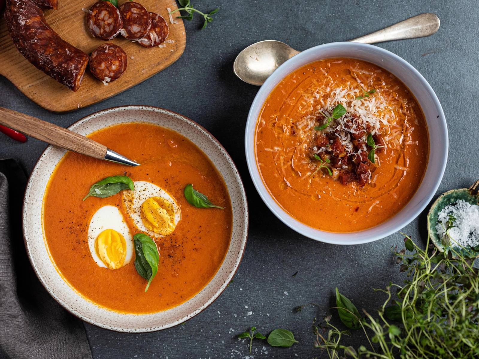 To skåler med tomatsuppe, en med egg og basilikum, og en annen med kjøttdeig og urter, på en mørk bordflate med urter og kjøttboller i bakgrunnen.