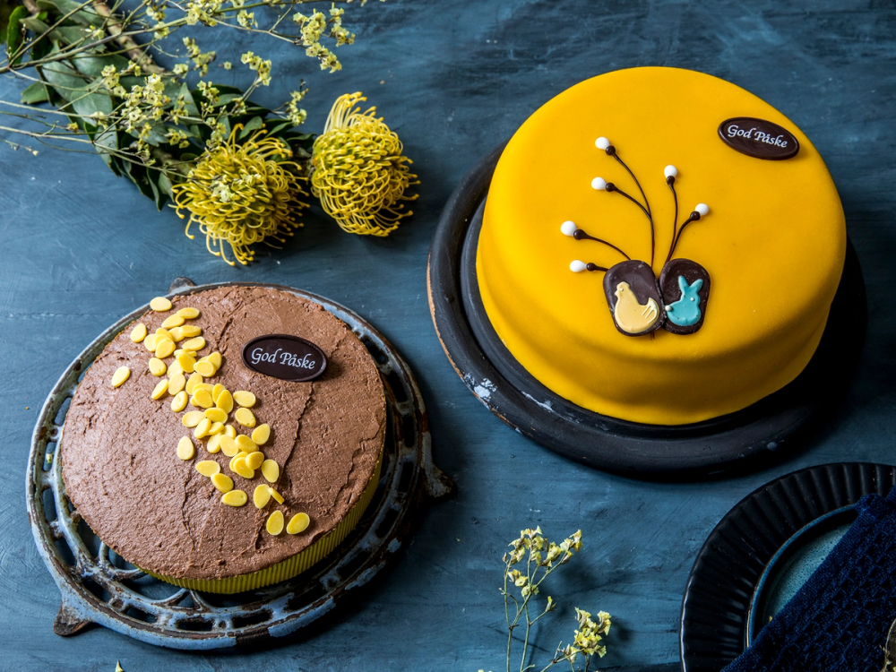 To kaker, en gul og en sjokoladekake, plassert på et bord med blomster rundt, for påske.