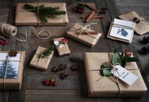 Pakkede julegaver med naturfargede papir, grønne grener, kanelstenger og julepynt på en mørk treoverflate.