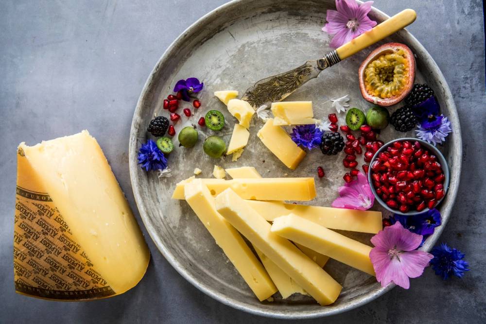 Ulike typer alpeost på en tallerken med bær, blomster og en ostehøvel, med en ostebit ved siden av.