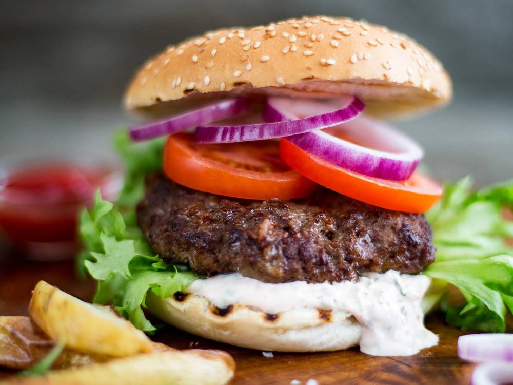En cheeseburger med revet ost, tomat, løk, salat og en kjøttkake i en sesambrødbolle, med pommes frites ved siden av.