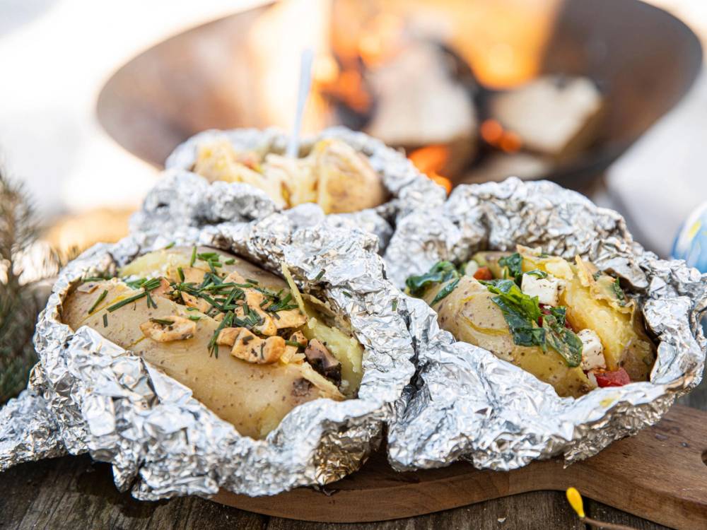 Bakt potet pakket inn i aluminiumsfolie, klar til å spises, med bål i bakgrunnen.