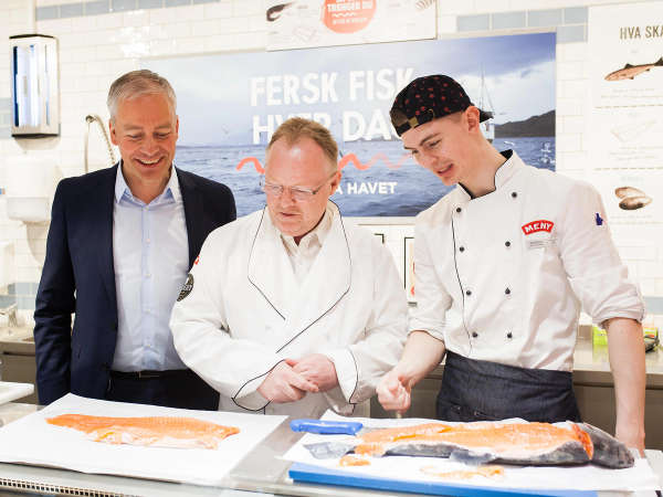 To personer i kokkeuniformer og en person i dress står bak en fiskebord i en butikk eller på et marked, med en stor fisk på bordet.