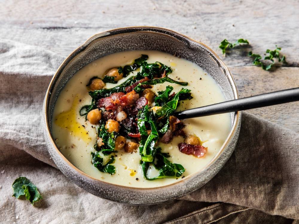 En skål med blomkålsuppe toppet med sprøstekt bacon, grønne urter og parmesanost, med en skje i suppen.