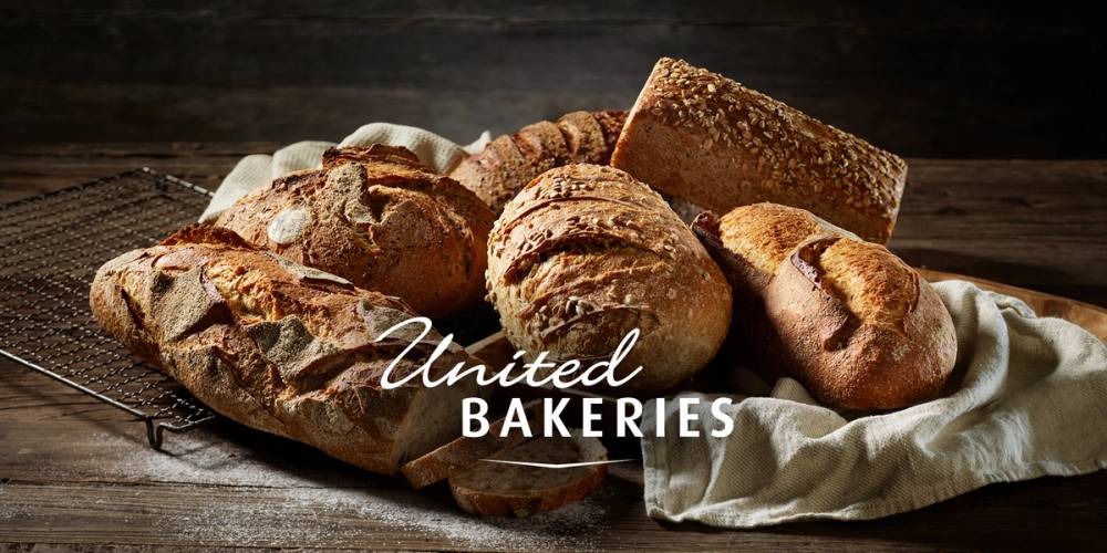 Et utvalg av ferskt bakst, inkludert brød og kaker, ligger på et brett med en klut under, med logoen 'United BAKERIES' over bildet.