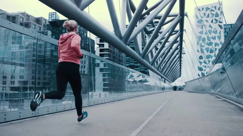 En person jogger på en moderne bro i en by med høye bygninger og en futuristisk arkitektur i bakgrunnen.