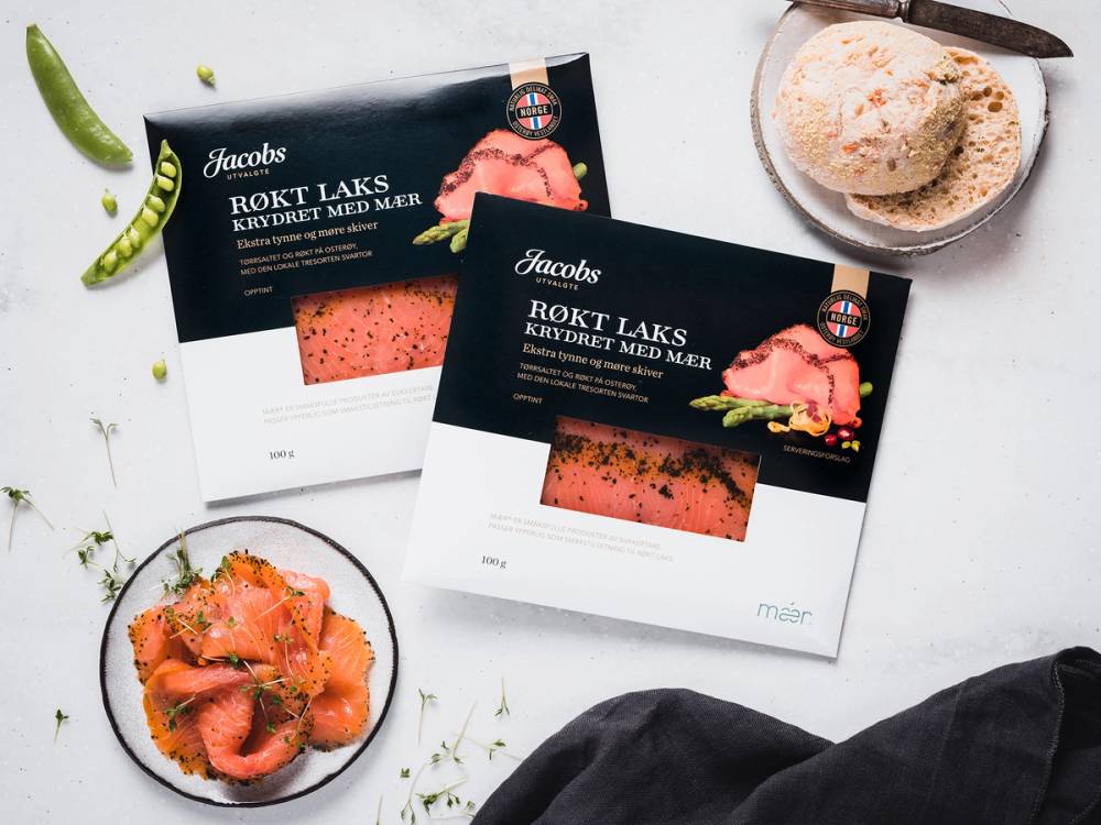 To pakker med Jacobs utvalgte røkt laks, en skål med skiver av røkt laks og en liten tallerken med en klatt smør eller krem, på et hvitt bord med noen grønne urter og limebiter.