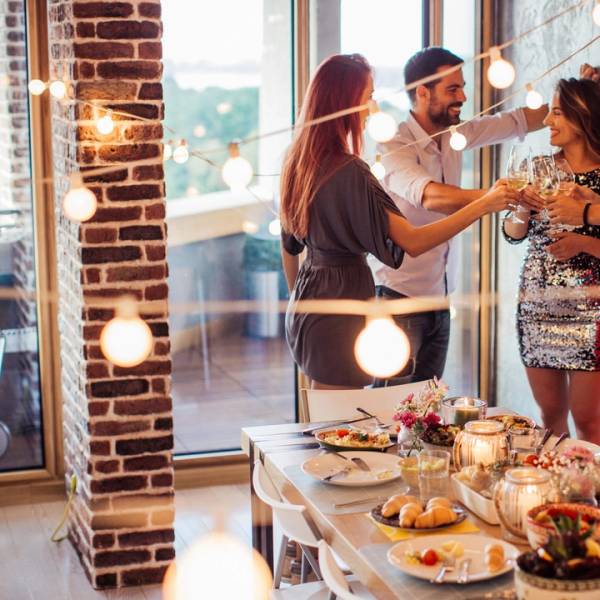 En gruppe venner skåler og har fest i et rom med store vinduer og eksponert murvegg, med en buffet av mat på bordet.