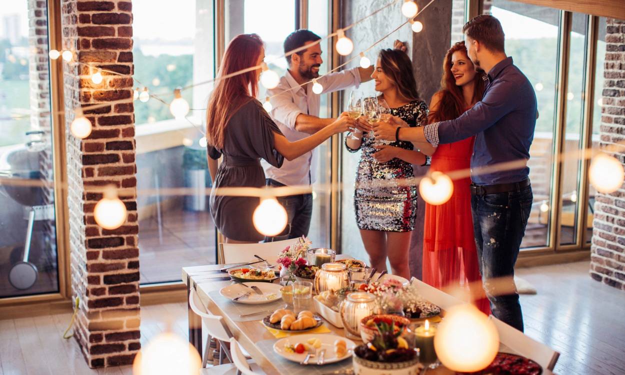 En gruppe venner skåler og har fest i et rom med store vinduer og eksponert murvegg, med en buffet av mat på bordet.