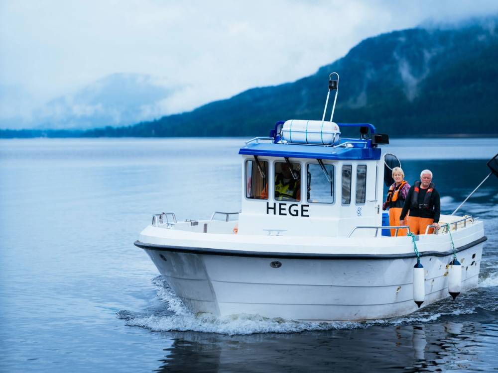 En hvit båttur på en rolig fjord med fjell i bakgrunnen, med to personer ombord, merket HEGE på båten.