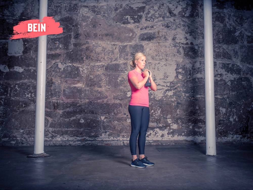 En kvinne står med hendene sammen foran brystet i et treningsrom med en steinvegg i bakgrunnen.