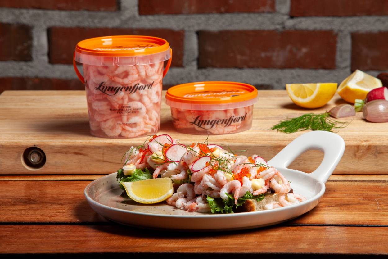 Reker på en tallerken med sitronbåter, urter og tilbehør, med to plastbokser med reker i bakgrunnen på et trebord.