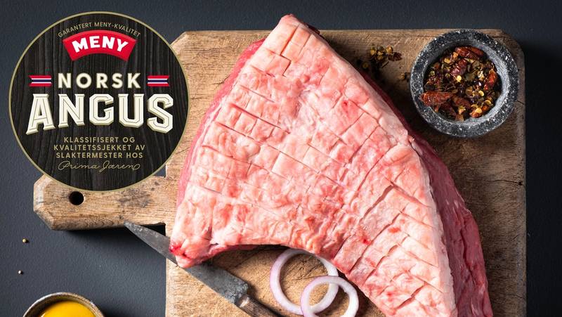 En stor rå Angus-biff med logo fra MENY på en skjærefjøl, omgitt av krydder og en liten skål med smør eller olje.