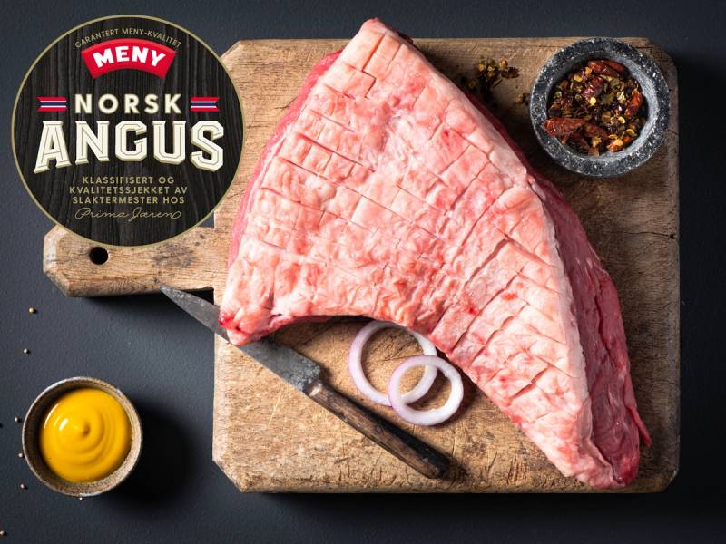 En stor rå Angus-biff med logo fra MENY på en skjærefjøl, omgitt av krydder og en liten skål med smør eller olje.