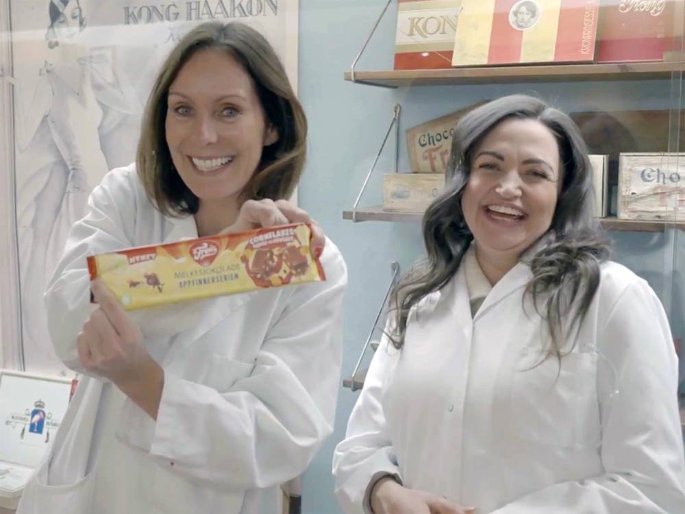 To personer i hvite labfrakker står i en butikk med Freia-sjokolade i hånden, og en butikkhylle med sjokoladeprodukter i bakgrunnen.