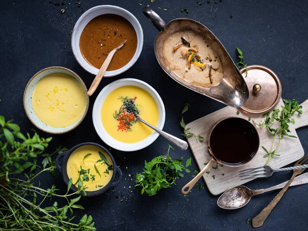 Ulike sauser i små skåler på et bord med urter og bestikk, sannsynligvis til matlaging eller servering.