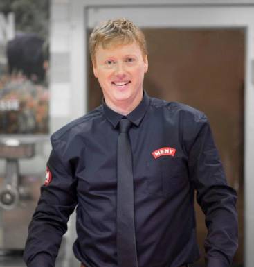 Vegard Kjuus i uniform med navnepatch, står foran en bakgrunn med industrielle elementer.