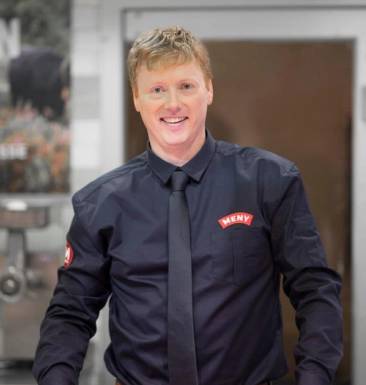 Vegard Kjuus i uniform med navnepatch, står foran en bakgrunn med industrielle elementer.