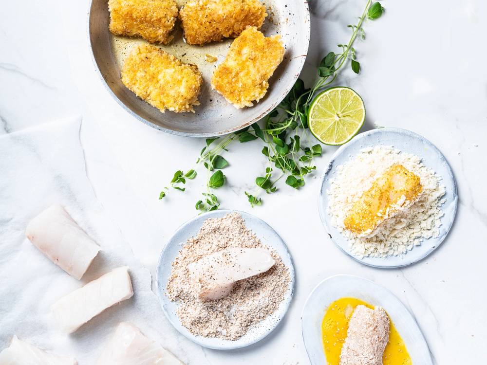 Fiskepinner på en tallerken med sitronbåter og urter, omgitt av mel og brødsmuler på et hvitt bord.