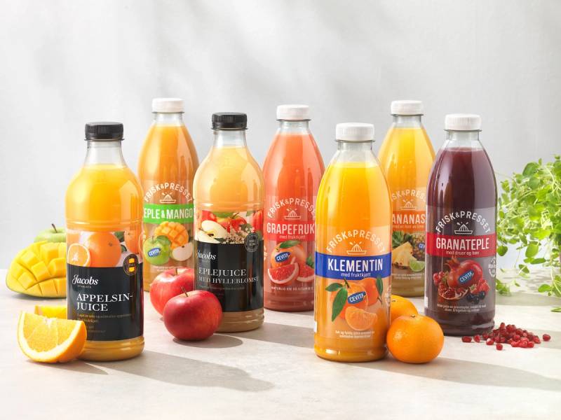 Seks ulike flasker med fruktjuice står på et hvitt bord, omgitt av frukt og berskiver, med en hvit bakgrunn.