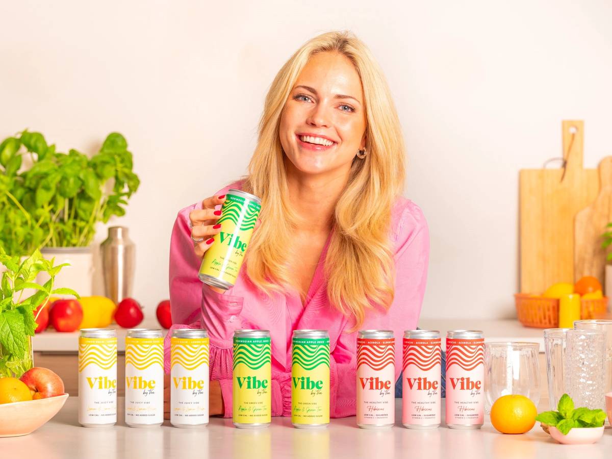 En person med lang blond hår som holder en Vibe By Voe-energidrikk i hånden, omgitt av flere bokser av samme merke på kjøkkenbenken.