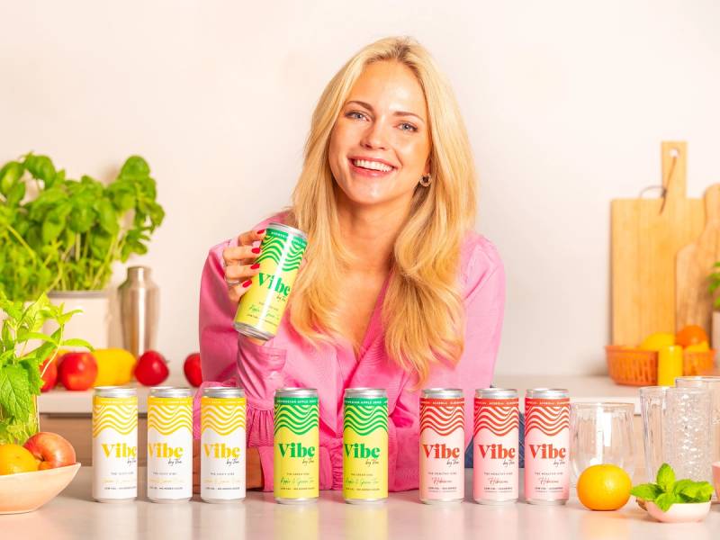 En person med lang blond hår som holder en Vibe By Voe-energidrikk i hånden, omgitt av flere bokser av samme merke på kjøkkenbenken.