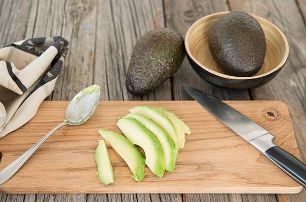 Avocado skrelles og kuttes på et skjærebrett med en kniv og skje, med to hele avokadoer og en skål med avokadobiter i bakgrunnen.